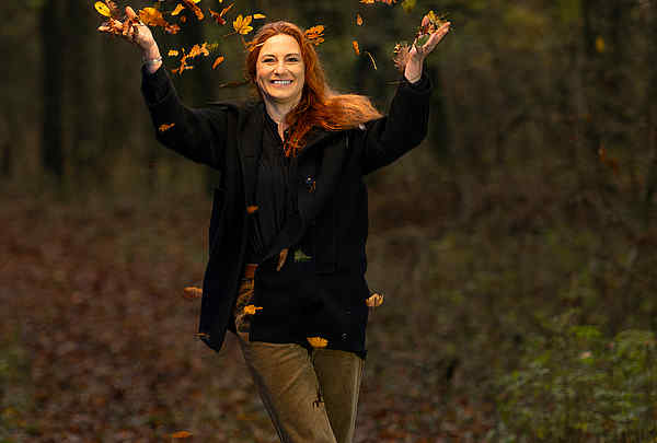 Frau mit roten Haaren und schwarzen Mantel steht im Wald und wirft Laub in die Luft