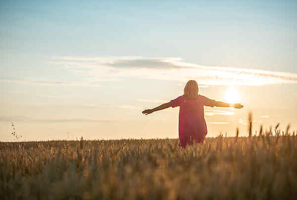 Frau im Kornfeld bei Sonnenuntergang mit magenta farbenen Kleid, breitet die Arme aus