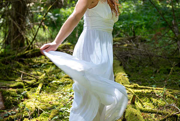 Fee mit roten Haaren und weißen Kleid steht im Wald auf dem moosbedeckten Boden im mystischen Licht und wedelt mit dem Kleid