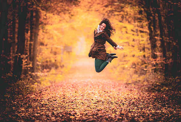 Junge Frau mit schwarzen Haaren springt im Laub, im Wald und lacht glücklich.