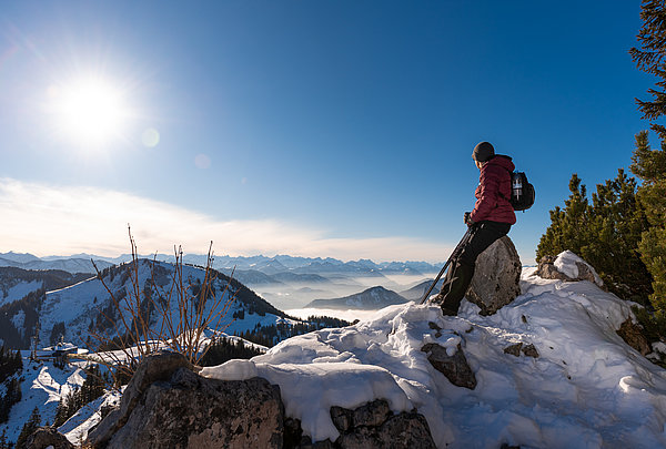 Junger Mann mit Bart auf dem Gipfel des Wallberg am Tegernsee mit okay Sonnenbrille und graue Wollmütze und rotem Anorak. Man schaut in die Sonne bei schneebedeckten Gipfel.