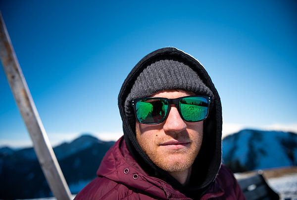 Junger Mann mit Bart, im Winter, auf dem Gipfel des Wallberg am Tegernsee mit okay Sonnenbrille und graue Wollmütze und rotem Anorak