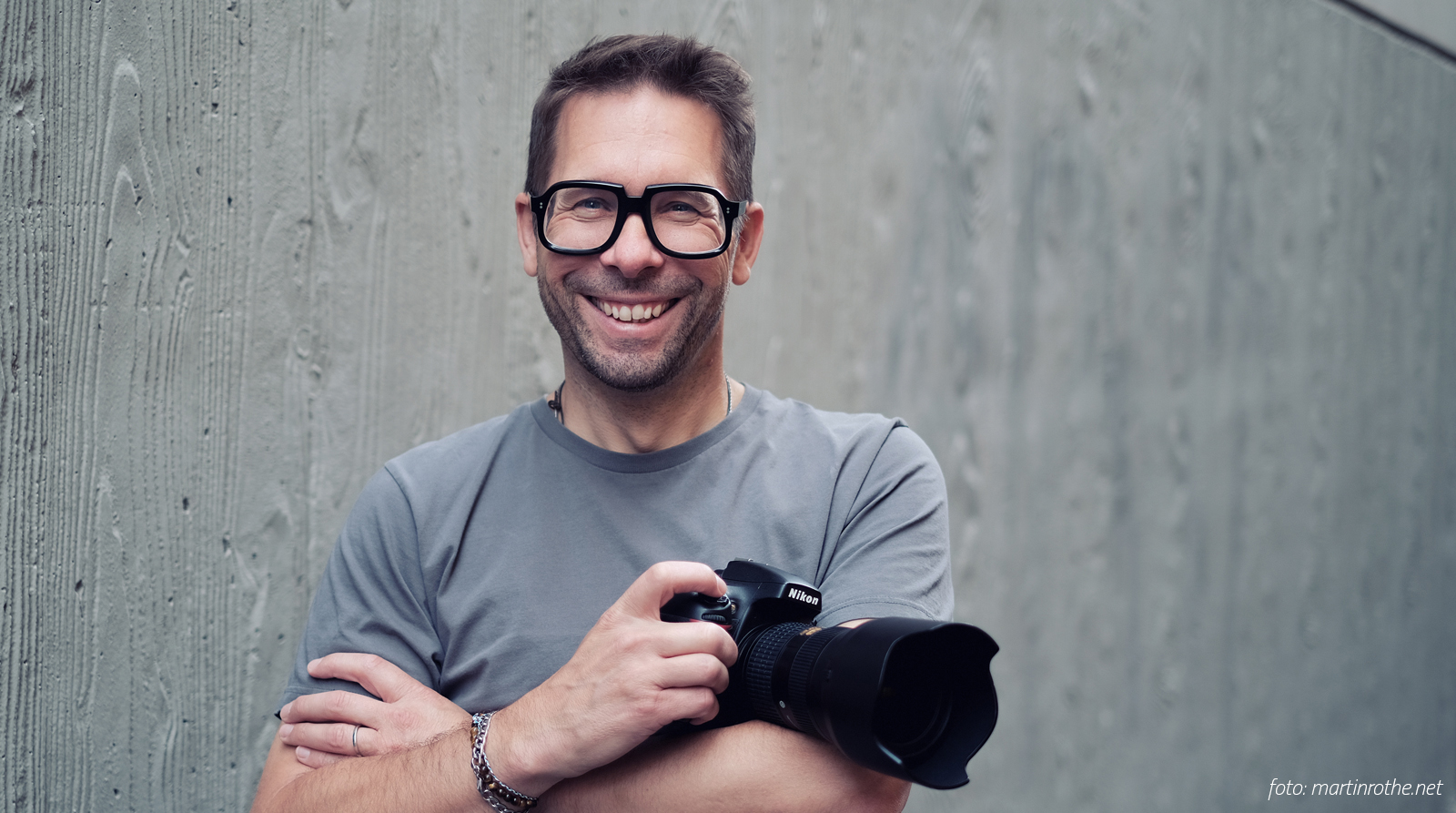 André Weber mit Kamera vor grauer Wand und stylishe Brille von Ottica Urbani